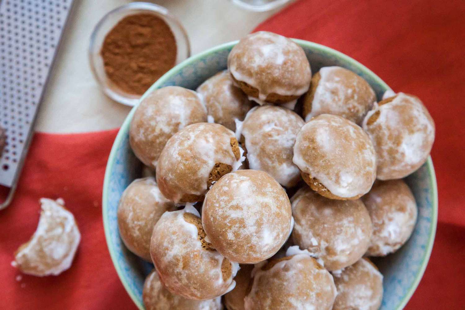 Pile of Pfeffernüsse Spice Cookies in a Bowl on a Red Table Cloth with One Cookie Bitten into Next to the Bowl. Surrounding the Bowl, a Small Bowl of Spice and a Grater.
