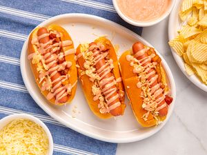 Three hot dogs on a plate with toppings and sauces accompanied by a side of chips and shredded cheese