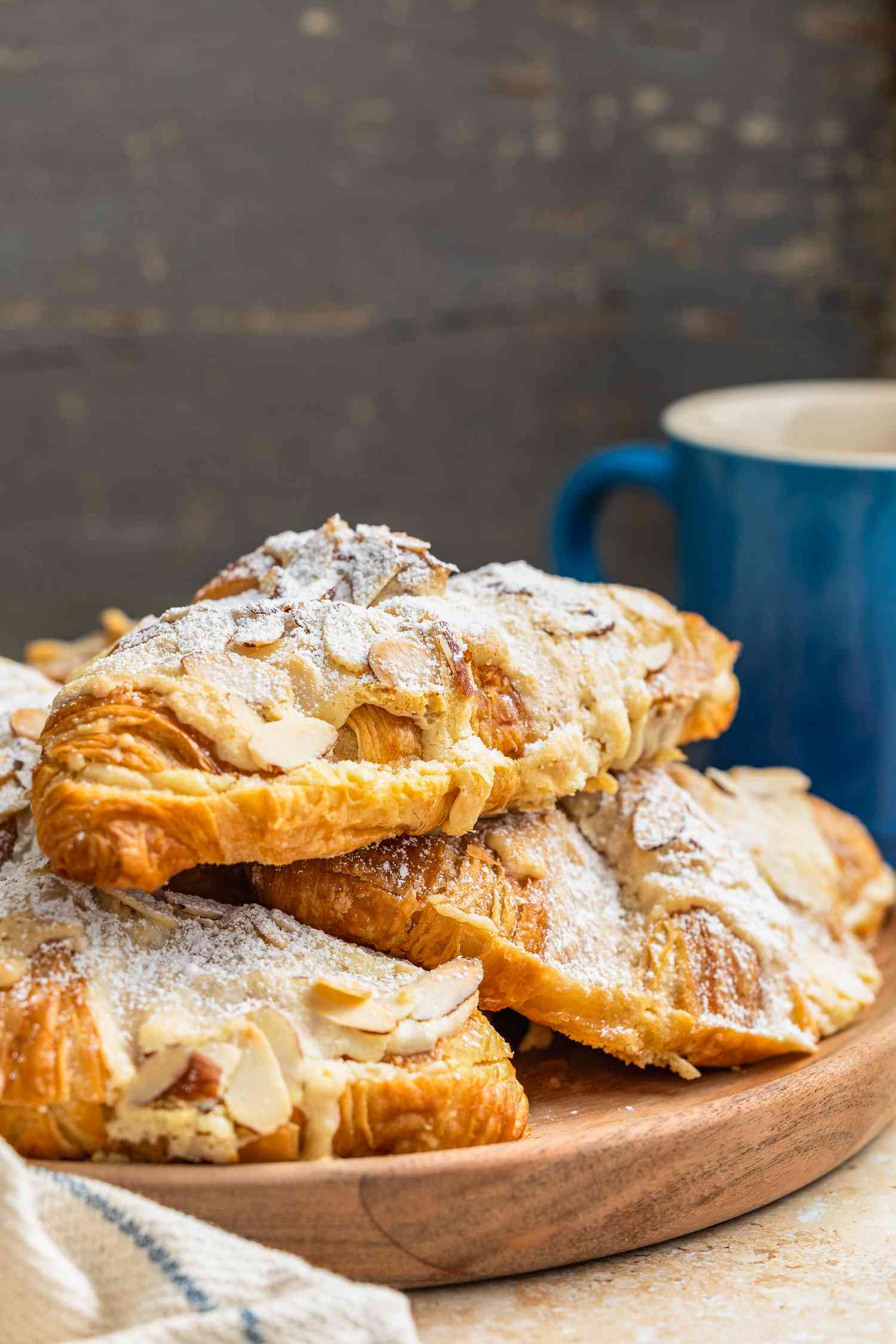 Pile of Almond Croissants on a Wooden Tray, and in the Surroundings, a Kitchen Towel and a Blue Coffee Mug