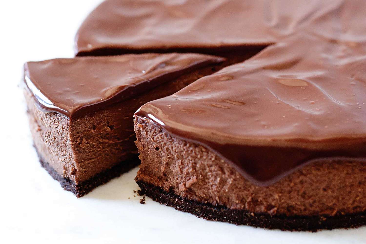 A slice taken out of chocolate cheesecake with dripping ganache frosting on a white background.