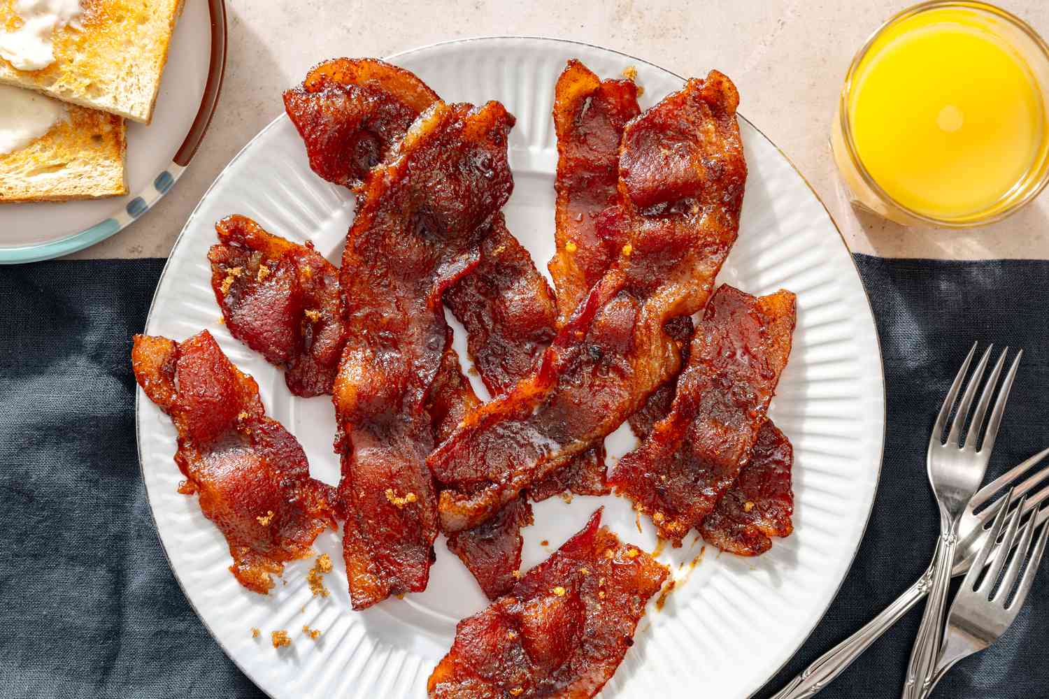Overhead view of a white plate of cooked bacon next to a glass of orange juice, a plate of toast and three forks