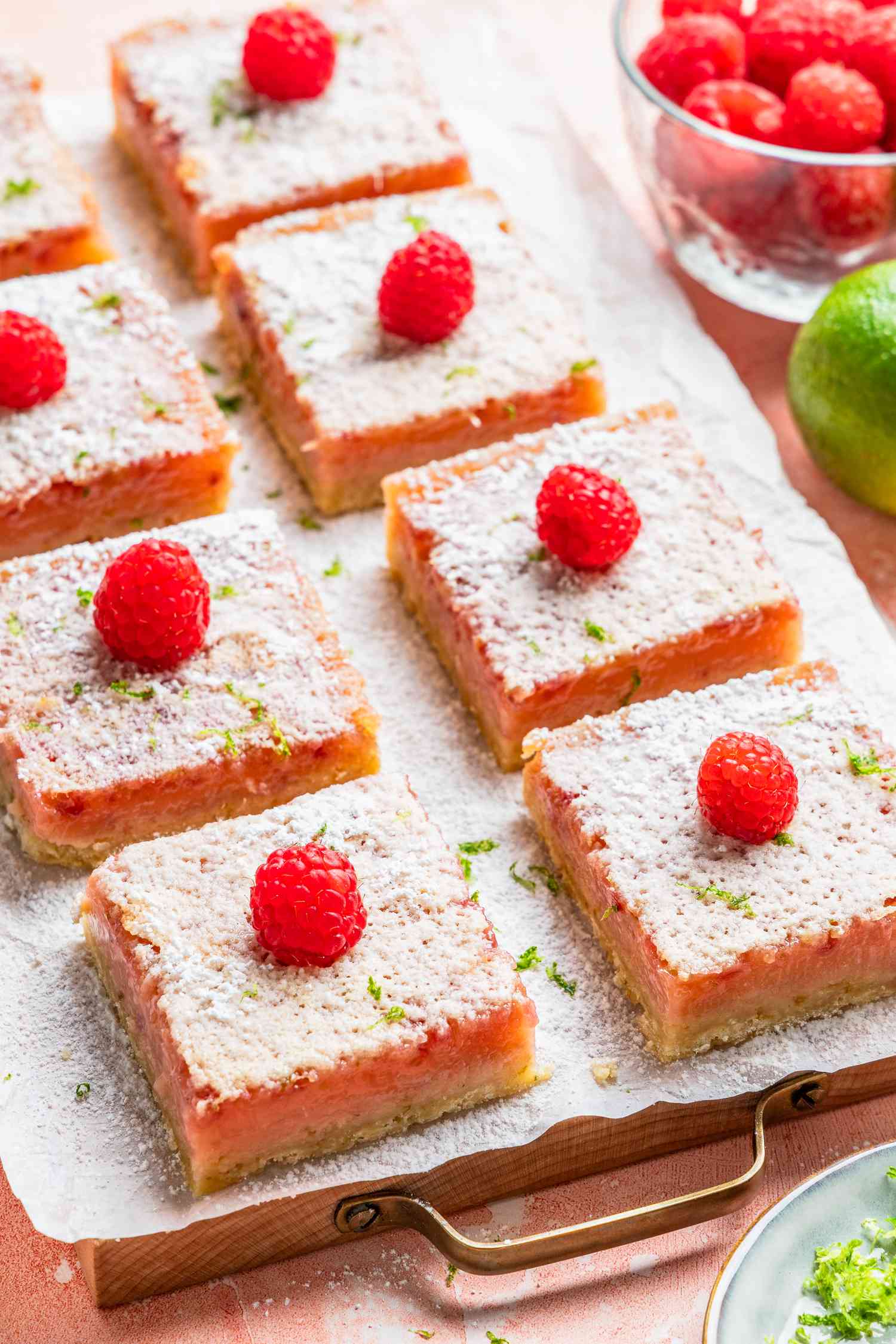 raspberry lime rickey bars on a parchment covered tray at a table setting with a jar of raspberries, a lime, and a small plate with lime zest 
