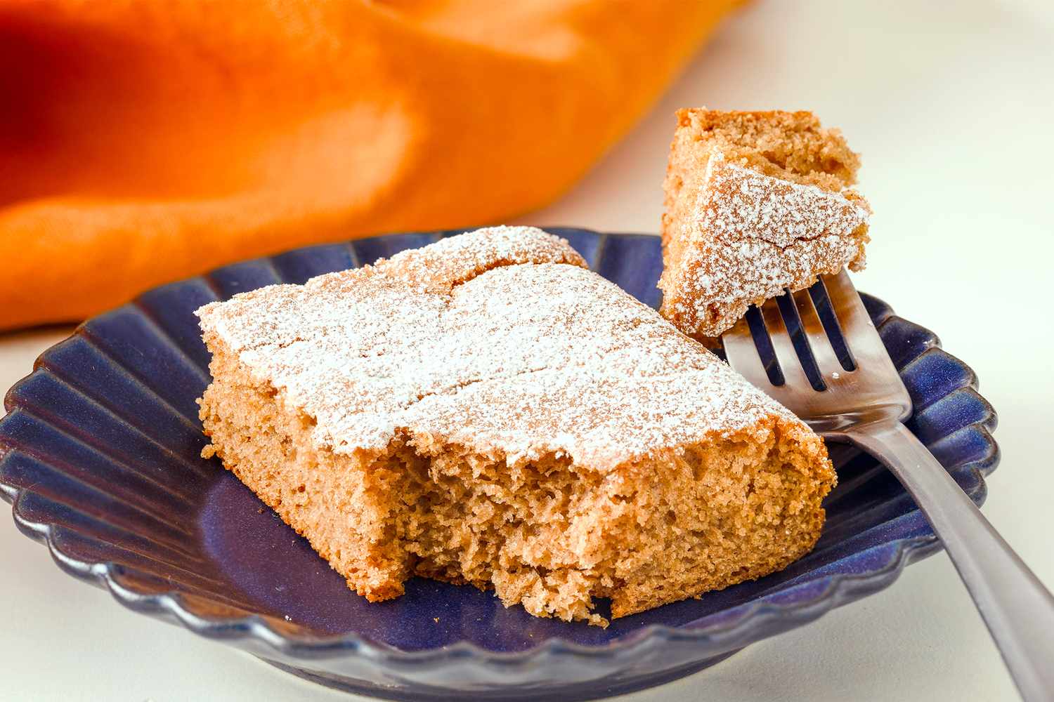 A slice of soda cake with powdered sugar on a plate with a fork cutting a piece