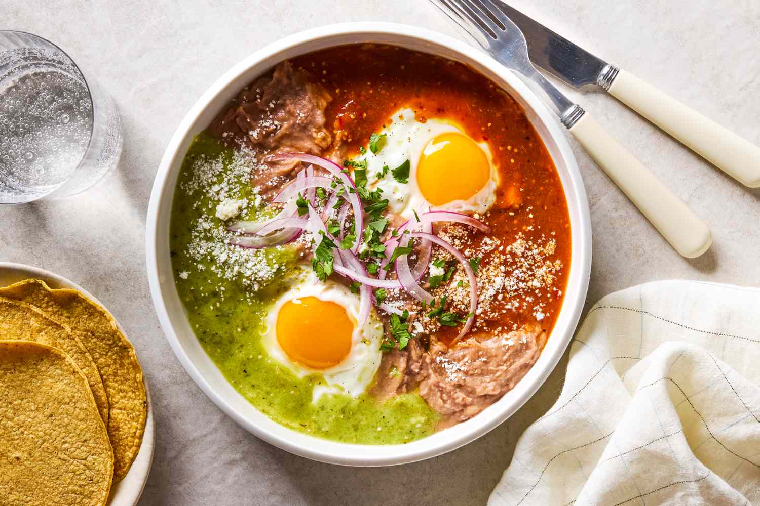A white bowl with Huevos Divorciados, next to a plate of corn tortillas and a glass of sparkling water