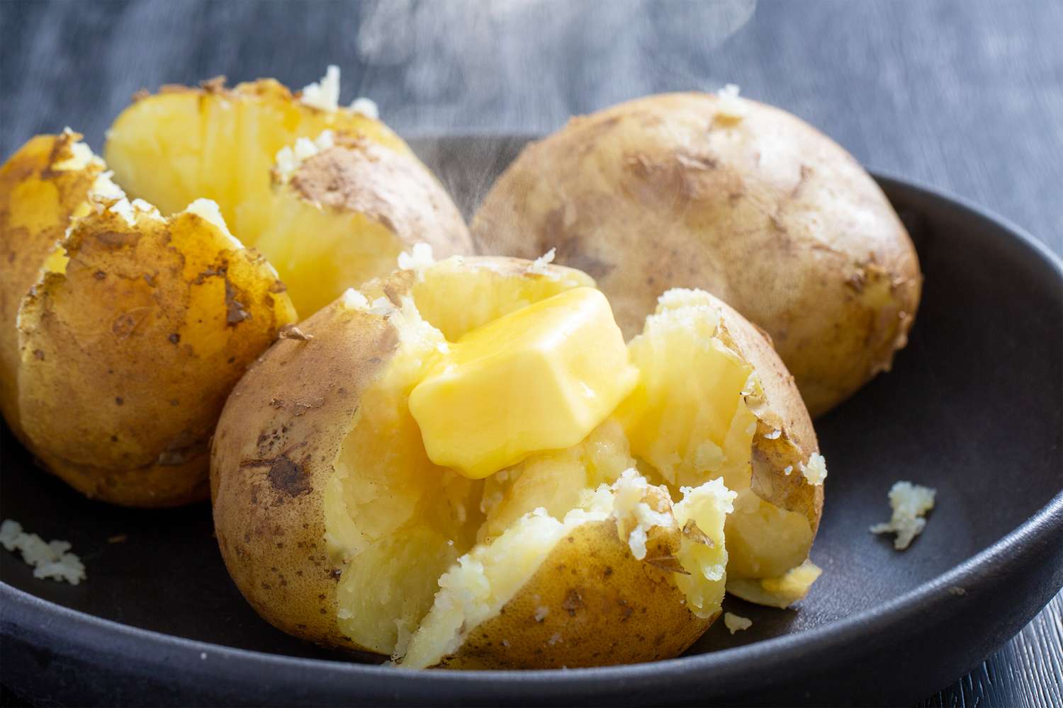 Three baked potatoes one cut open with butter melting on top served on a dark plate