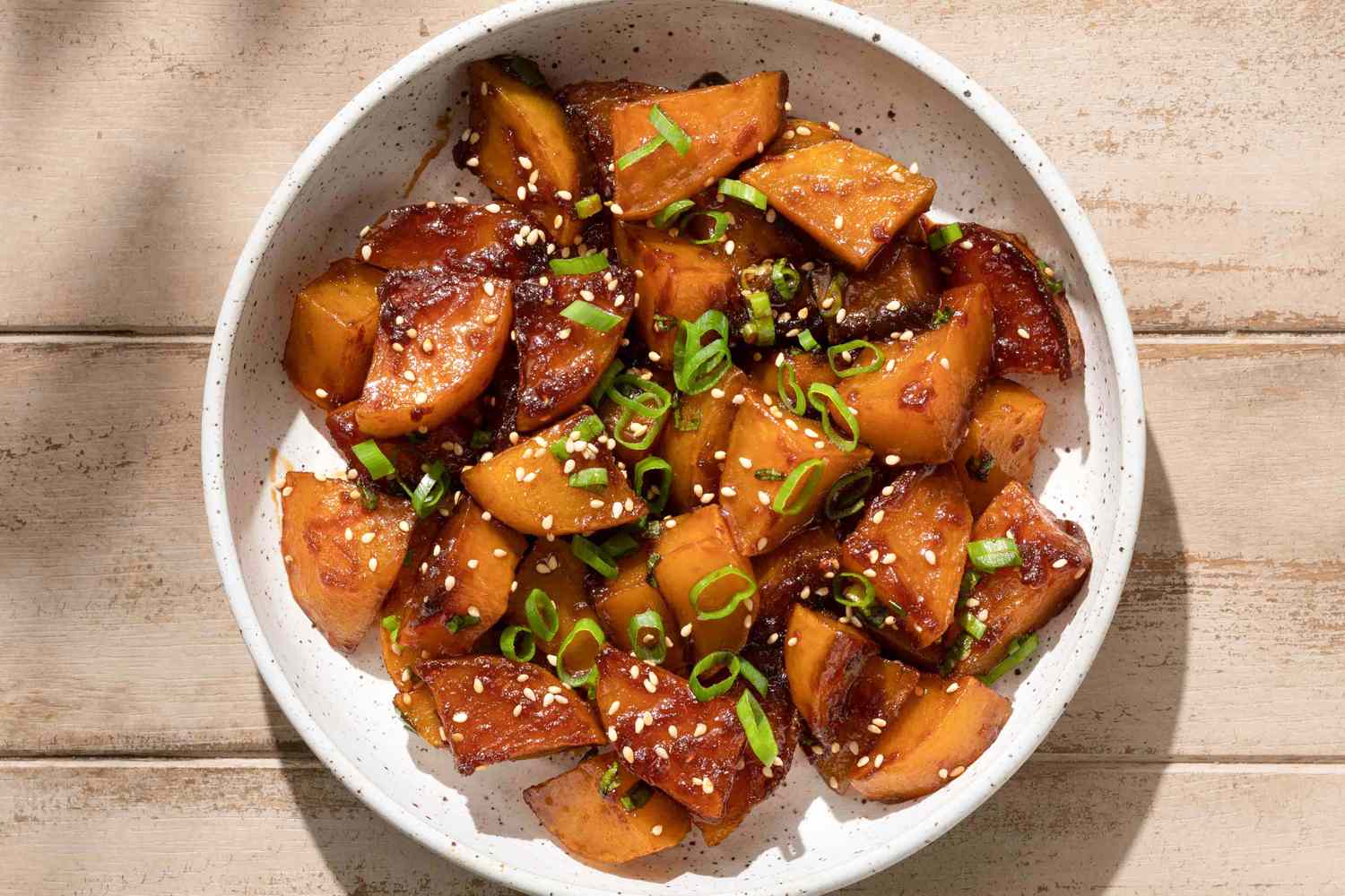 Overhead view of a white bowl of glazed potato slices topped with sesame and scallions for Gamja Jorim (Korean Braised Potatoes) recipe