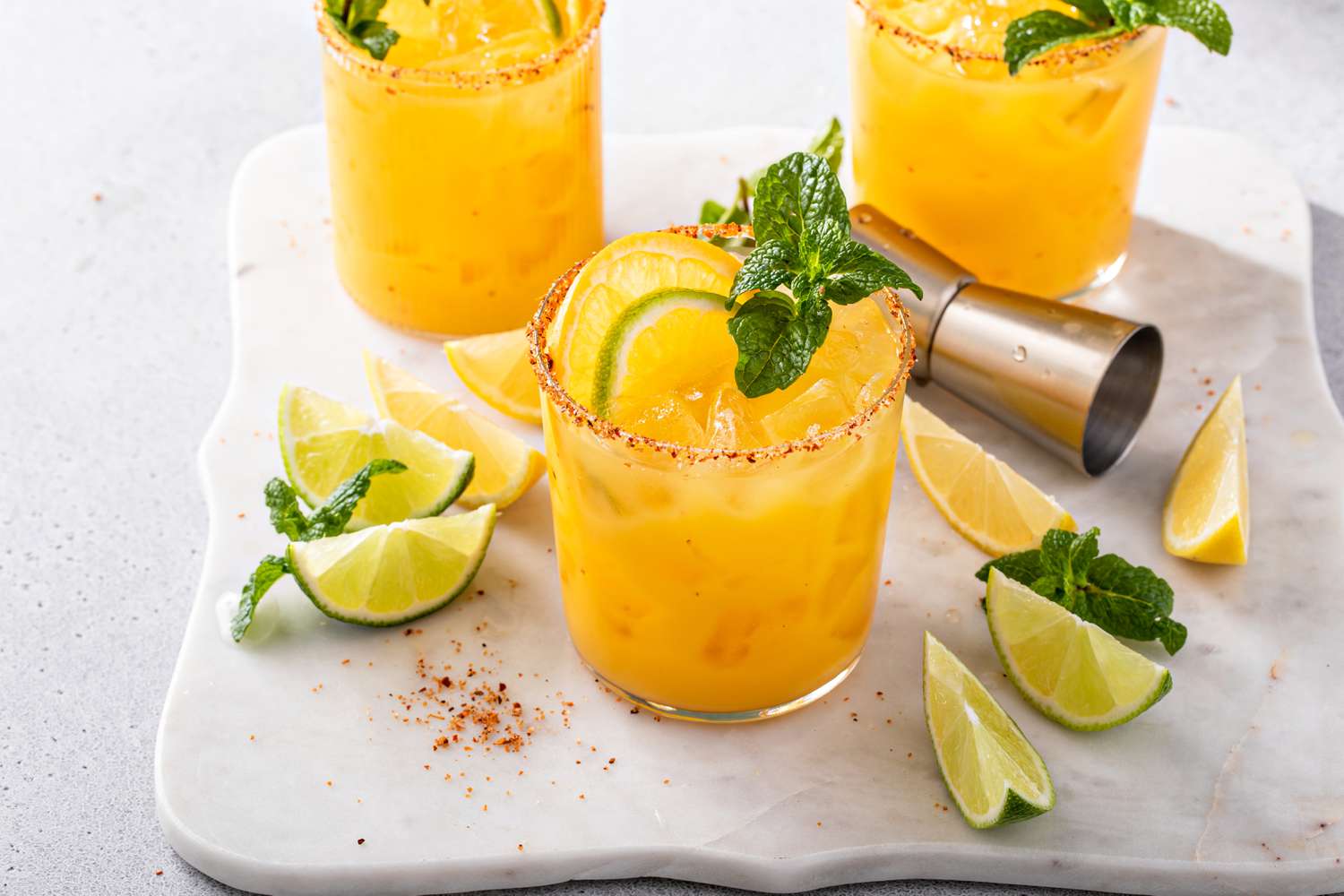 margaritas in three glasses (garnished with lime slices and mint sprigs), lime wedges, and a cocktail measuring cup all on a marble tray
