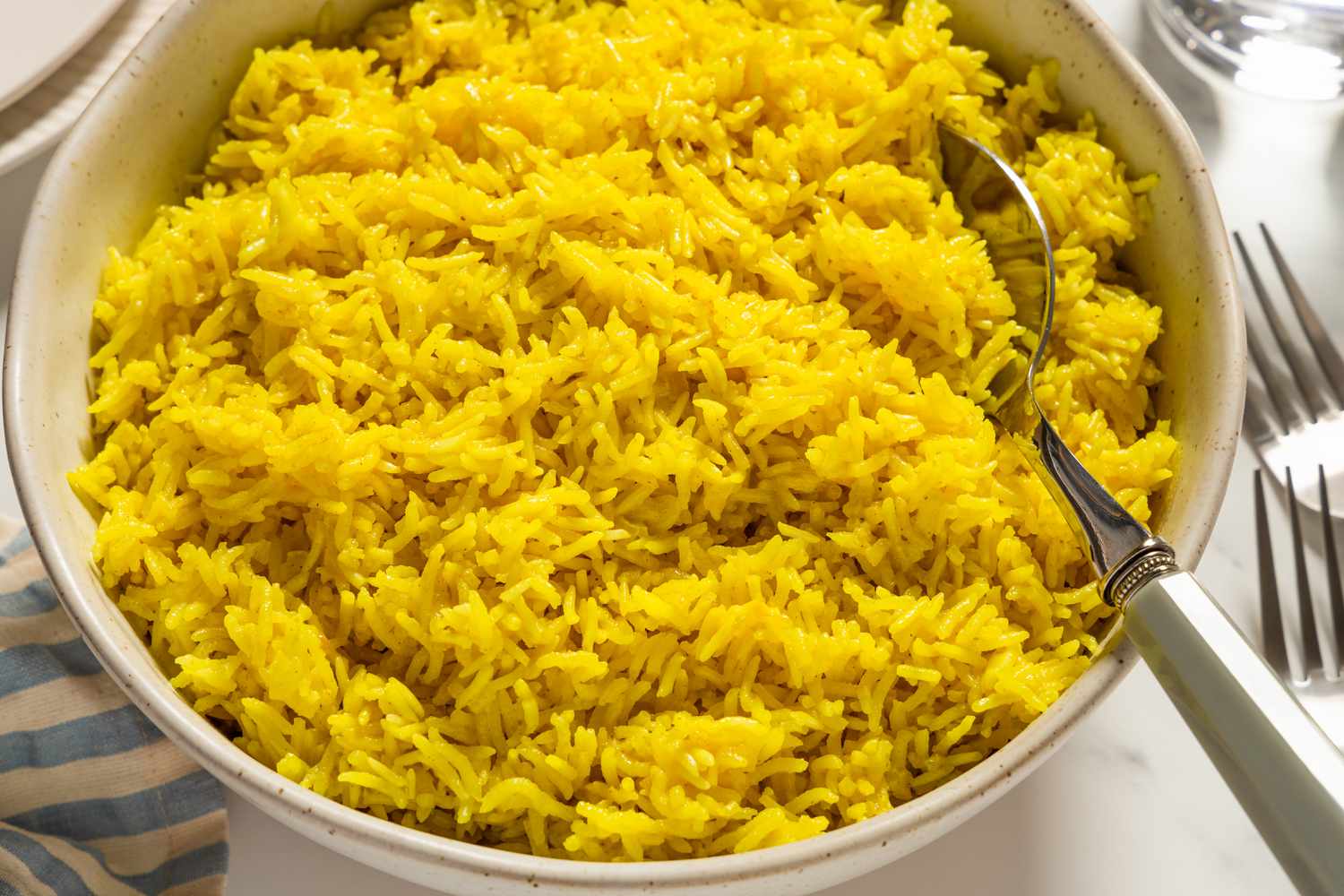 Closeup view of a large bowl of golden rice with a serving spoon all on marble countertop with a cloth napkin, forks, plates a glasses