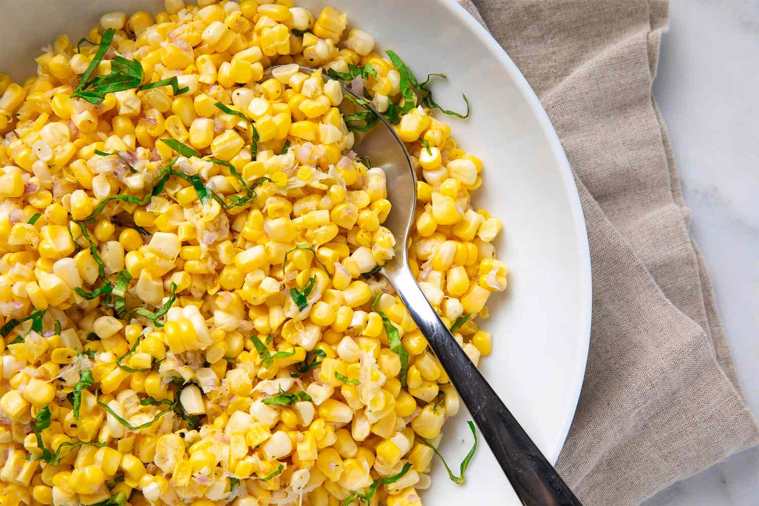 A bowl of corn salad garnished with herbs a serving spoon positioned in the bowl