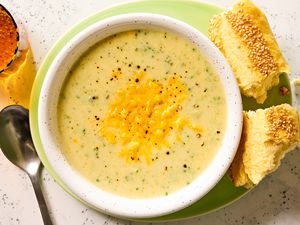 Overhead view of a bowl of broccoli cheddar soup on a plate with pieces of bread and next to a spoon and drink glass