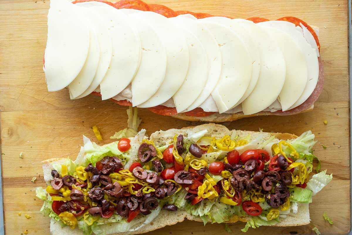 Italian club sandwich with meat and cheese layered on one half and lettuce, tomato, peppercinis and olives layered on the other half.