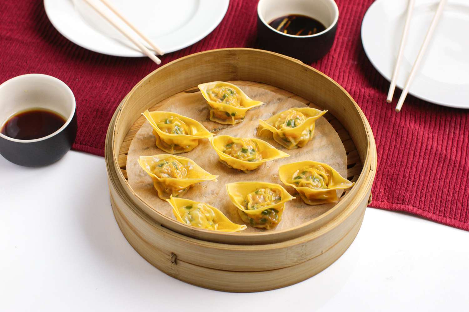 Pork and Chive Dumplings in a Lined Bamboo Steamer Next to a Kitchen Towel with Two Bowls of Vinegar Sauce and Two Plates on Top