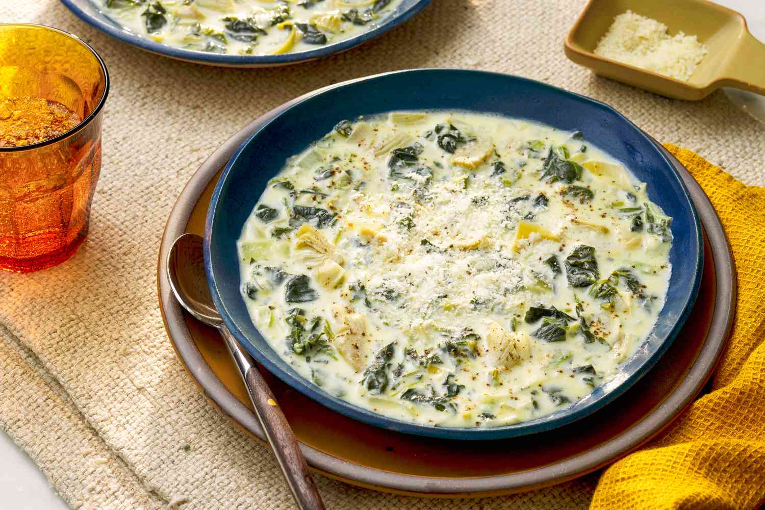 Bowl of spinach artichoke dip soup on a plate at a a table setting with another bowl of soup, a small dish of parmesan, a glass fo water, and mustard colored table napkins