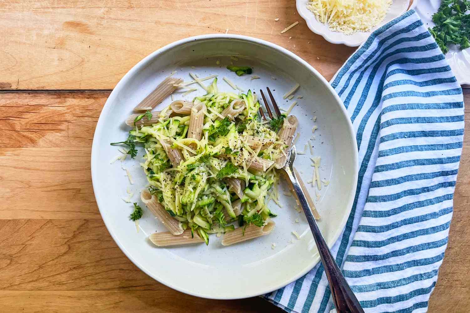 Plate of penne pasta with shredded zucchini and grated cheese fork placed on the side wooden surface and striped cloth visible nearby