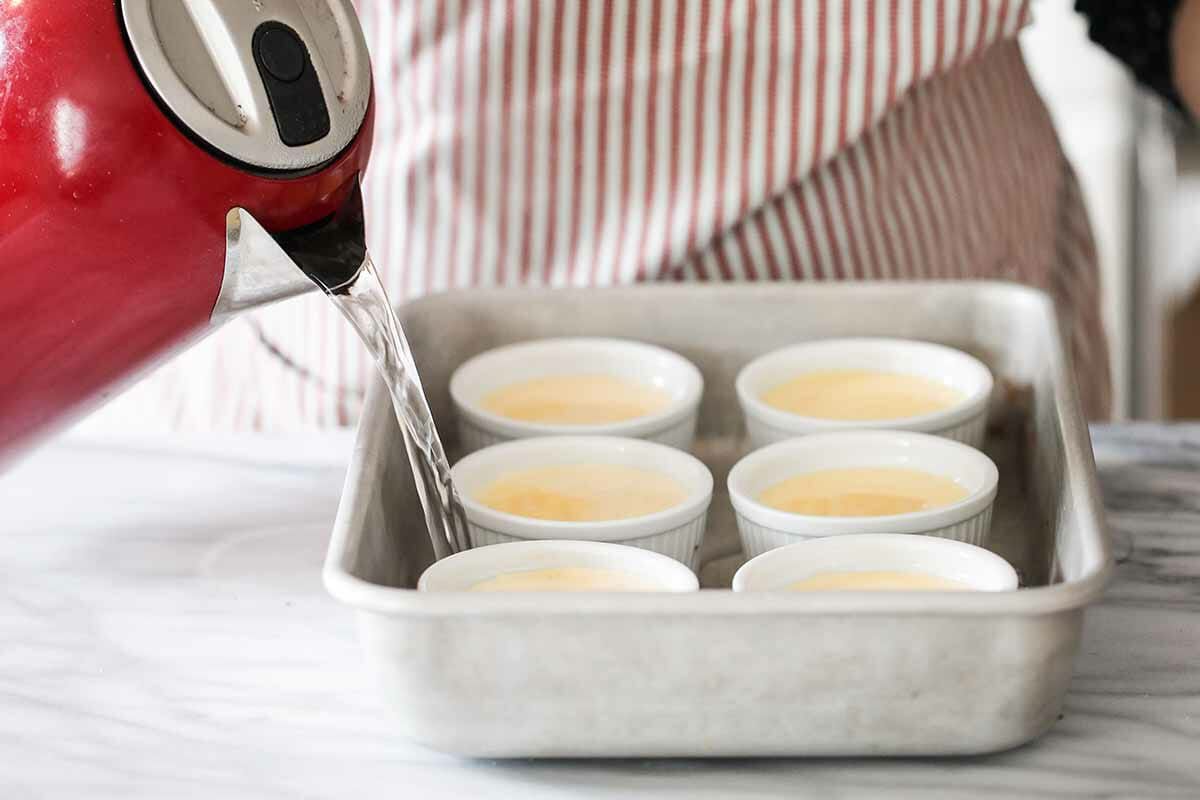 A hot water bath being poured in a baking dish to heat six ramekins 