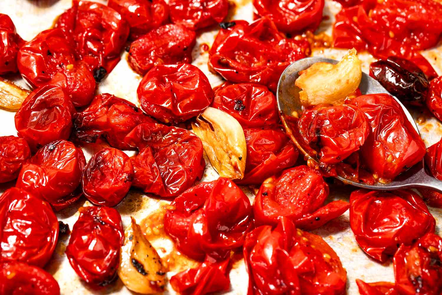 Blistered cherry tomatoes and garlic on a baking sheet with a spoon