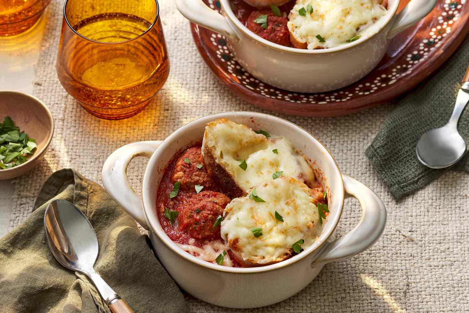 Meatball sub soup in a pot-like ramekin at a rustic table setting with another serving in a ramekin, spoons on table napkins, bowl of parsley, and glasses of water 
