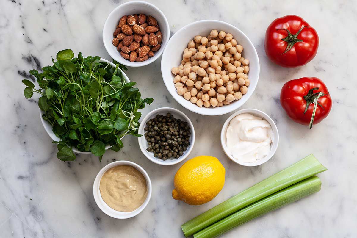 Ingredients for an easy sandwich recipe. Bowls include: almonds, canned chickpeas, capers, mustard, mayonnaise, sprouts, lemon, celery and tomatoes.
