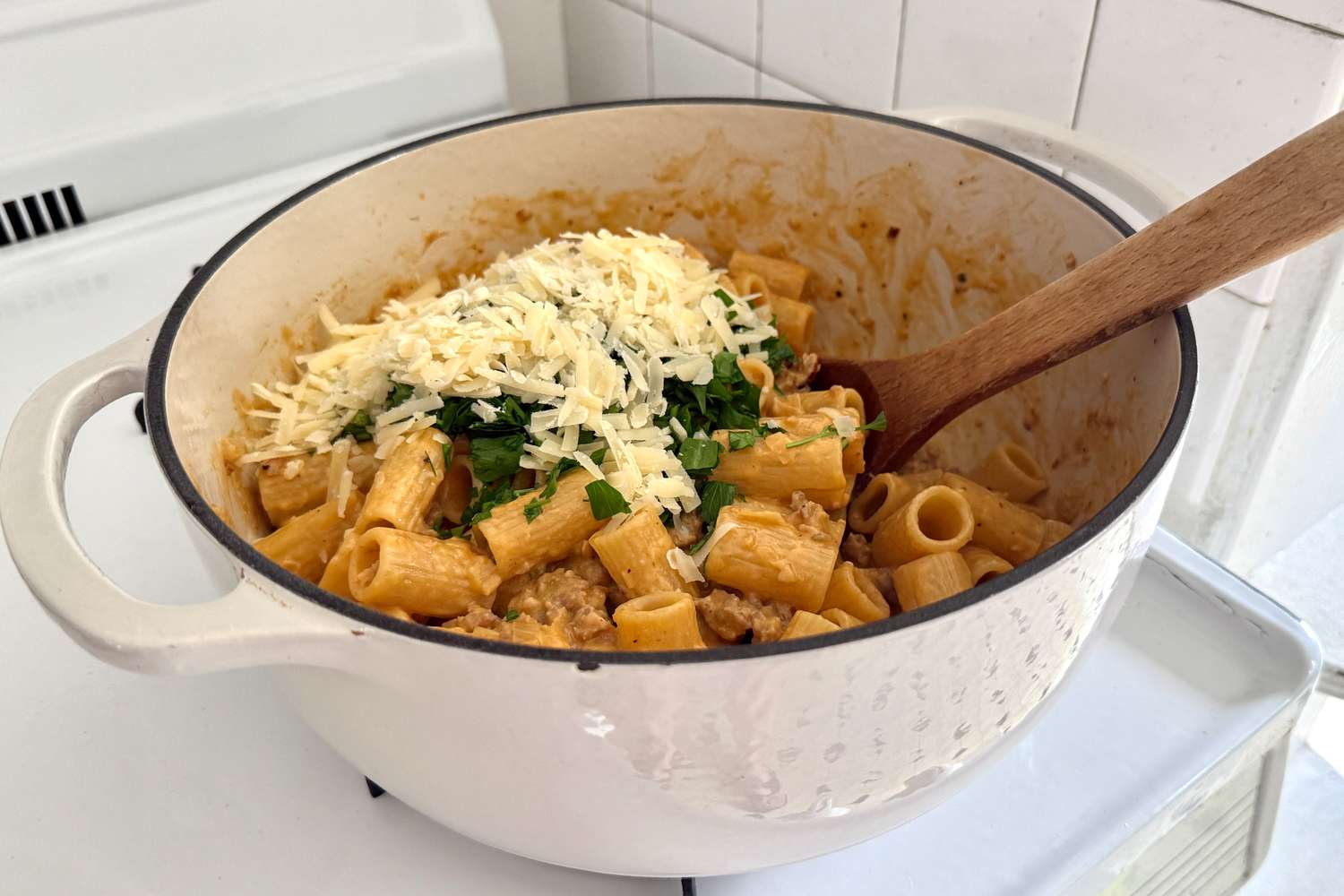A pot of rigatoni pasta topped with grated cheese and herbs with a wooden spoon placed inside