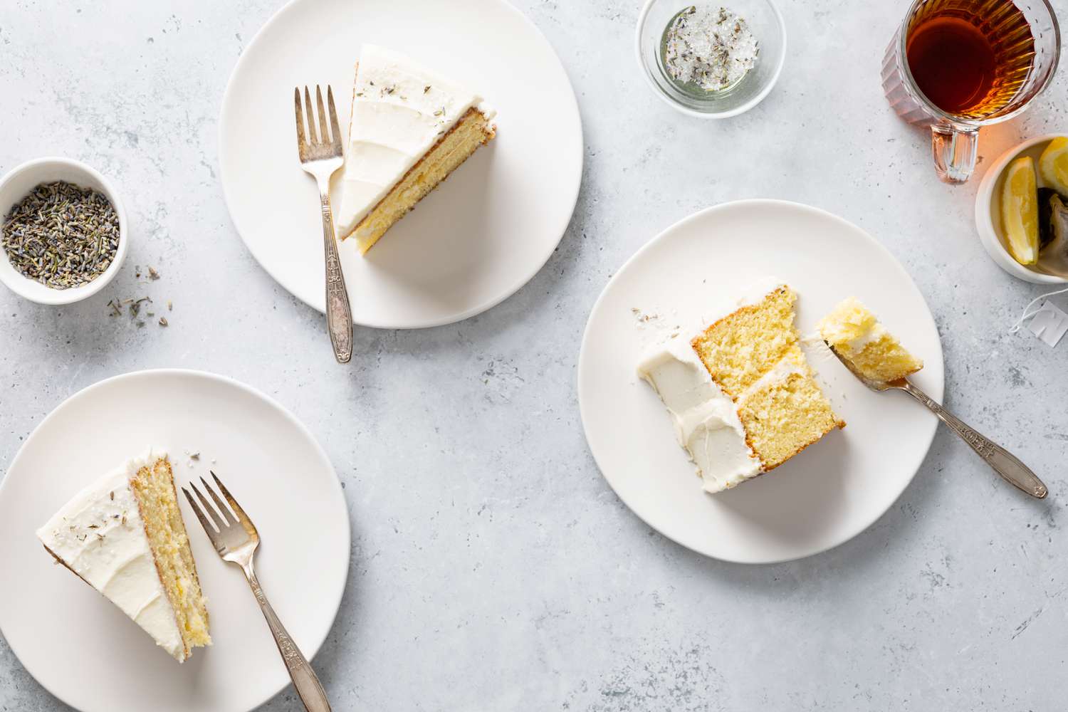 Three Slices of Lemon Lavender Cake on a Three Plates with a Fork Each, Next to a Cup of Tea, a Small Bowl of Lavender, and a Dish of Lemon 
