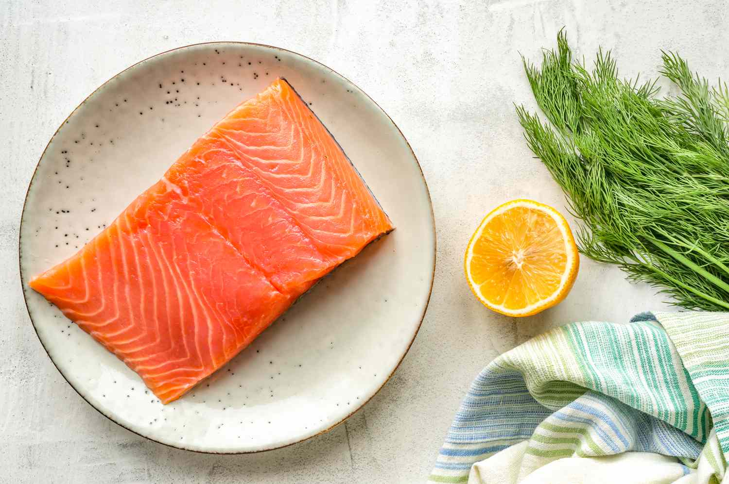 A bright pink fillet of raw salmon on a plate next to a lemon half and a bunch of dill
