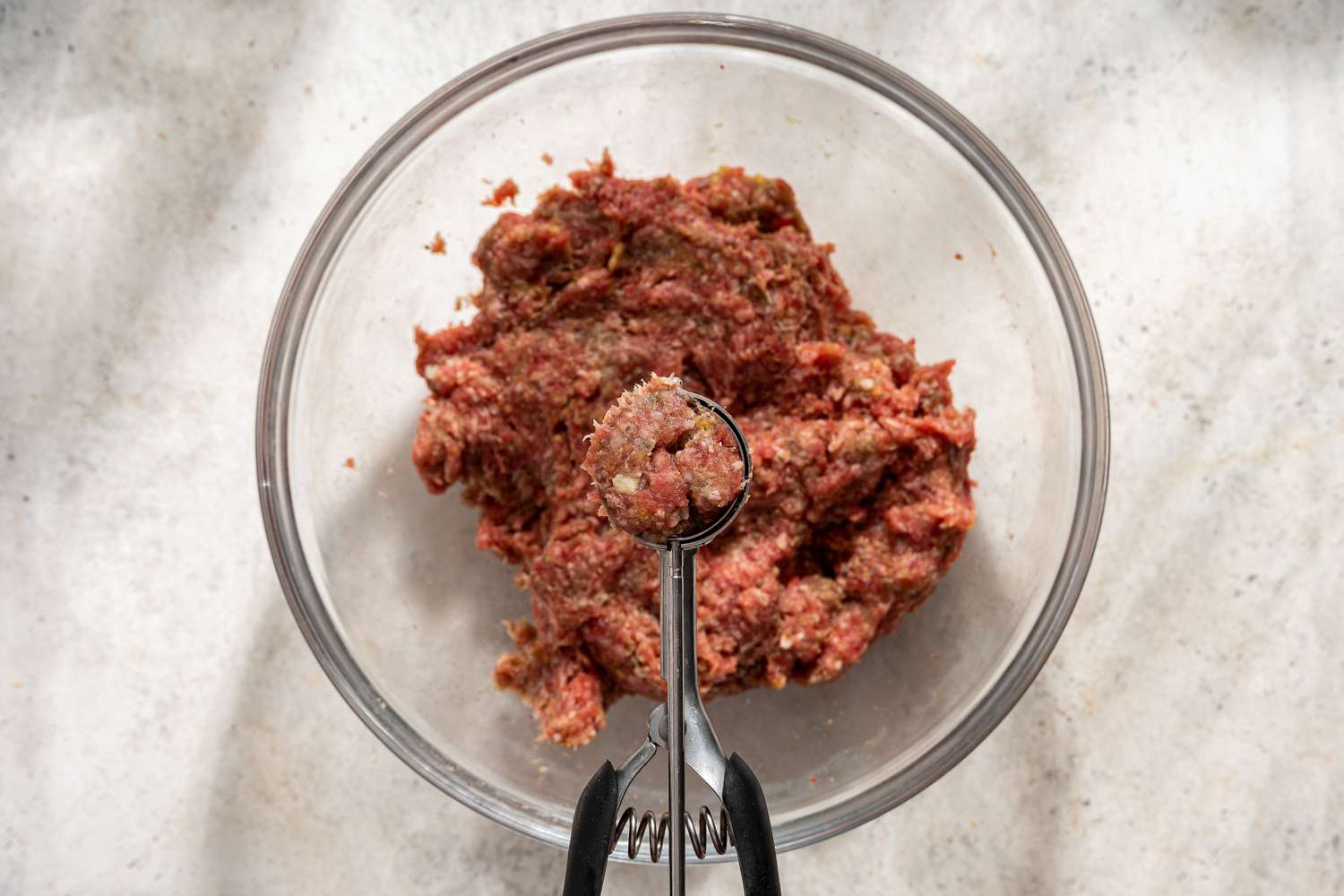 Overhead view of scooping ground beef mixture out of a clear glass bowl with a cookie scoop to make meatballs for Italian Meatball Soup recipe