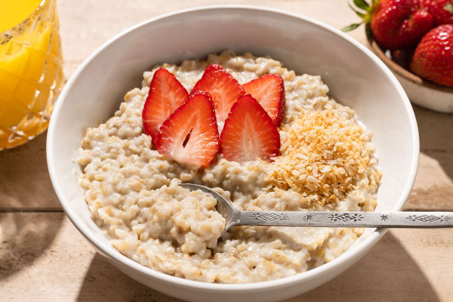 Bowl of oatmeal topped with sliced strawberries and shredded coconut, spoon resting inside, a glass of juice and fresh strawberries nearby.