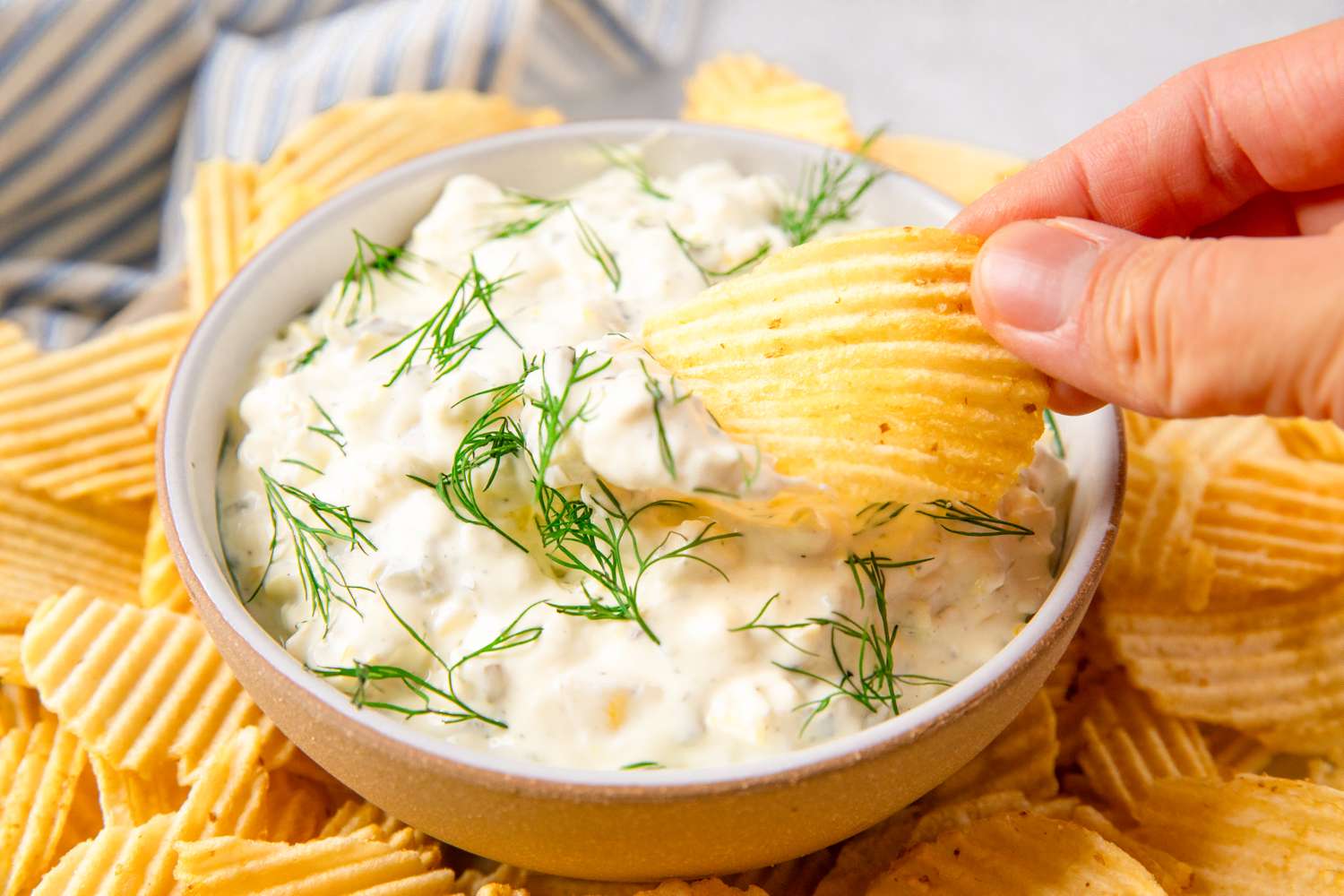 A bowl of creamy pickle dip garnished with fresh herbs, surrounded by crinkled chips, with a hand dipping one of the chips