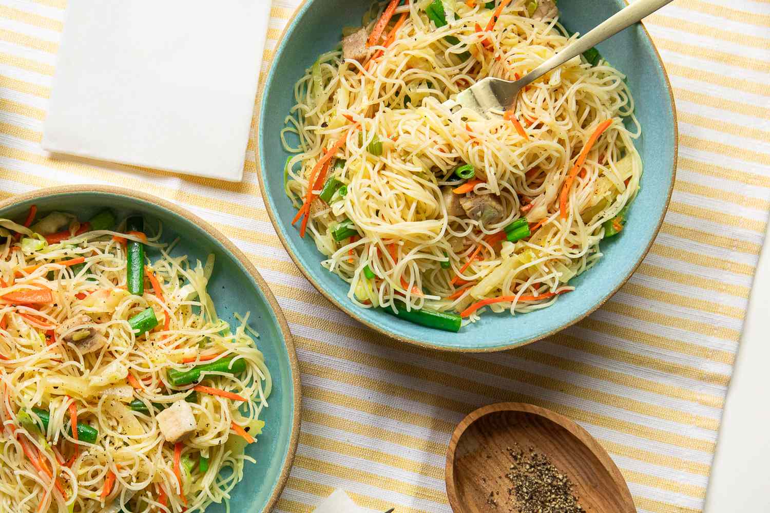Two Bowls with Pancit Bihon (Filipino Rice Noodles) on a Placemat Next to a Bowl of Cracked Pepper