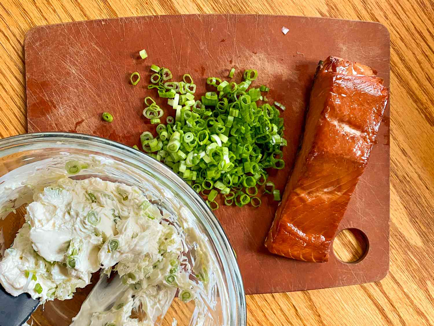 Overhead view of a cutting board on a wooden tabletop with chopped green onions and a piece of salmon next to a clear glass bowl of cream cheese being mixed with green onions for 3-ingredient smoked salmond dip