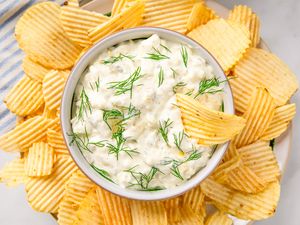 A bowl of pickle dip garnished with dill surrounded by potato chips arranged on a plate