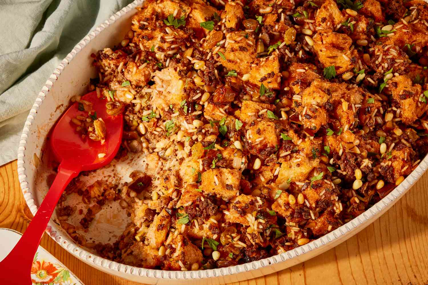 Greek-style stuffing in an oval white serving dish, with a red serving spoon in the right corner where some of the stuffing has been scooped out