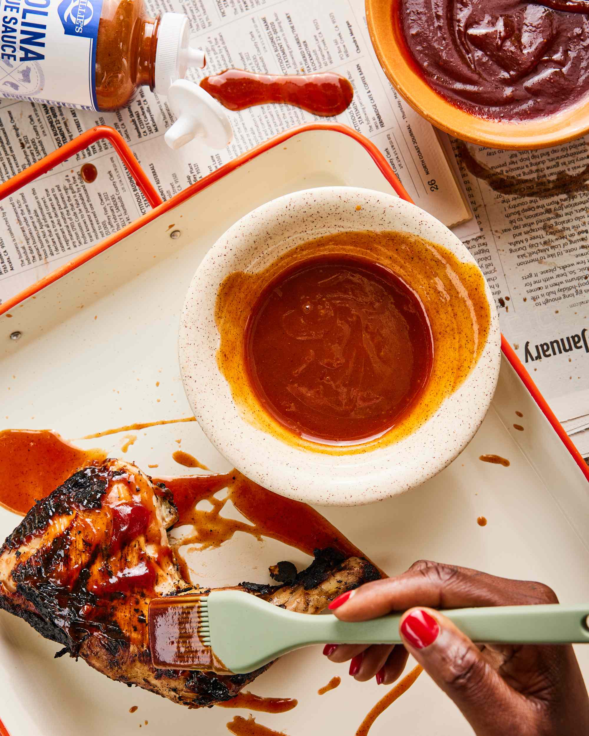 Sauce being brushed onto grilled chicken with a basting brush, barbecue sauce and a small bowl are in the scene