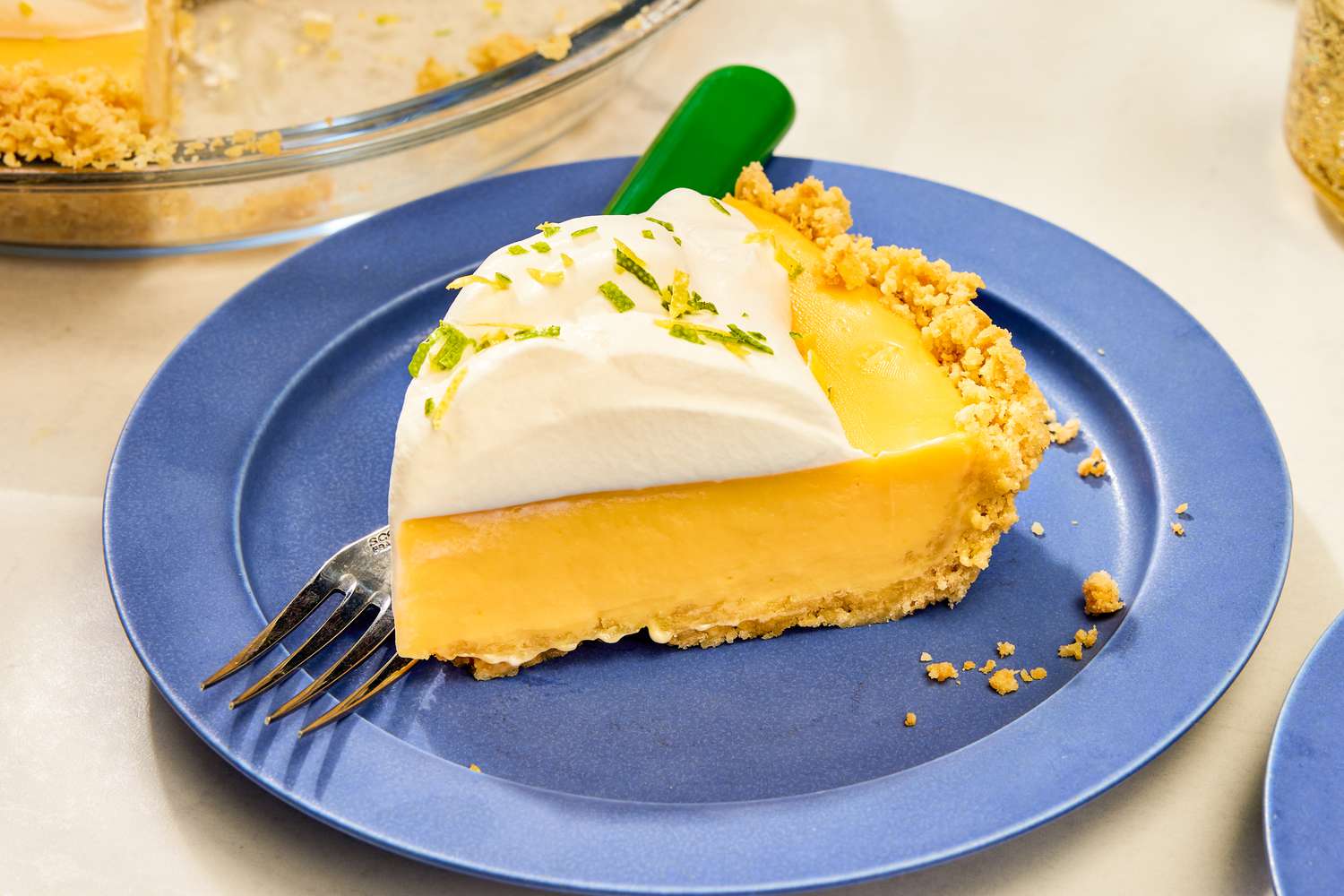 Slice of pie on a blue plate, topped with whipped cream and lime zest, with a fork on the plate