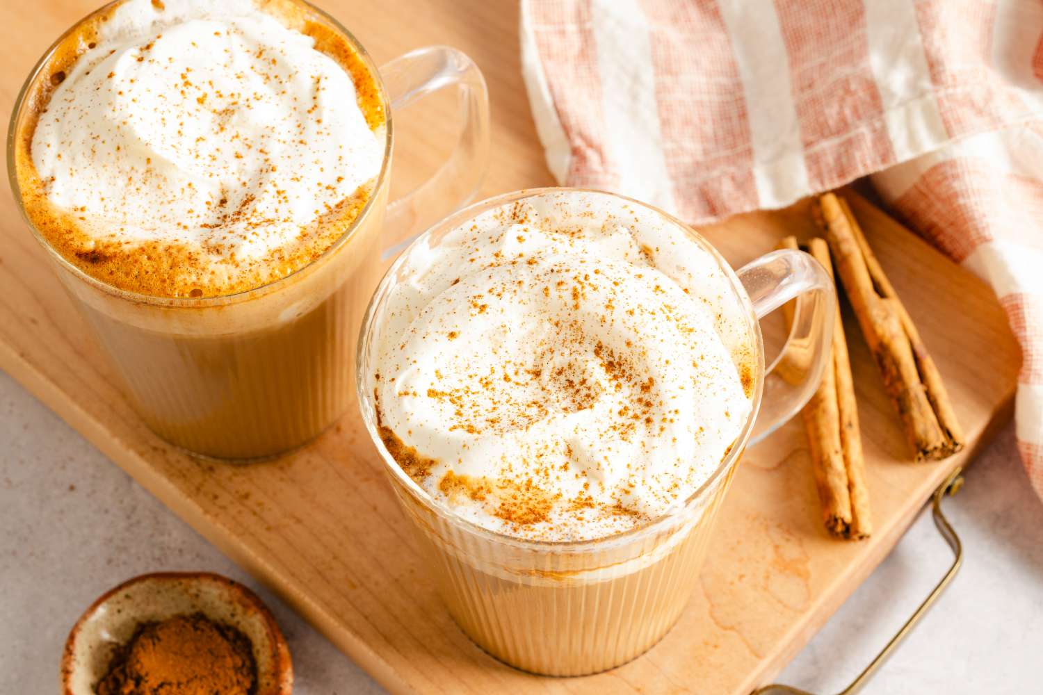 Two pumpkin spice lattes topped with whipped cream and ground spices on a wooden tray accompanied by cinnamon sticks and a cloth napkin