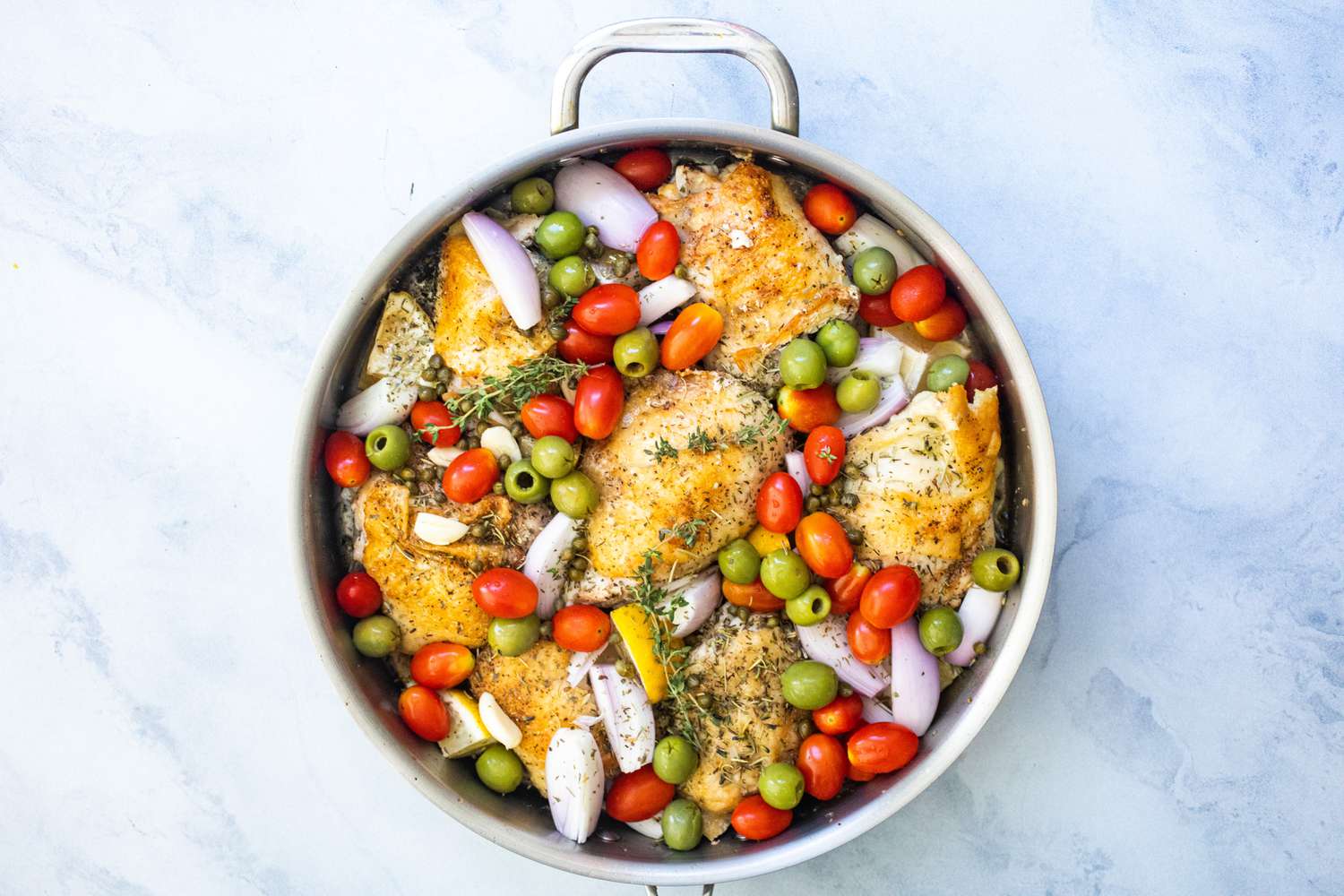 Chicken with shallots, garlic, cherry tomatoes, green olives, lemon wedges, capers, and thyme sprigs in pan