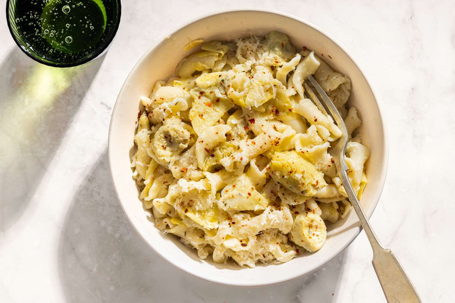 A bowl of pasta with artichokes lemon and seasonings served with a fork on a white surface beside a beverage glass