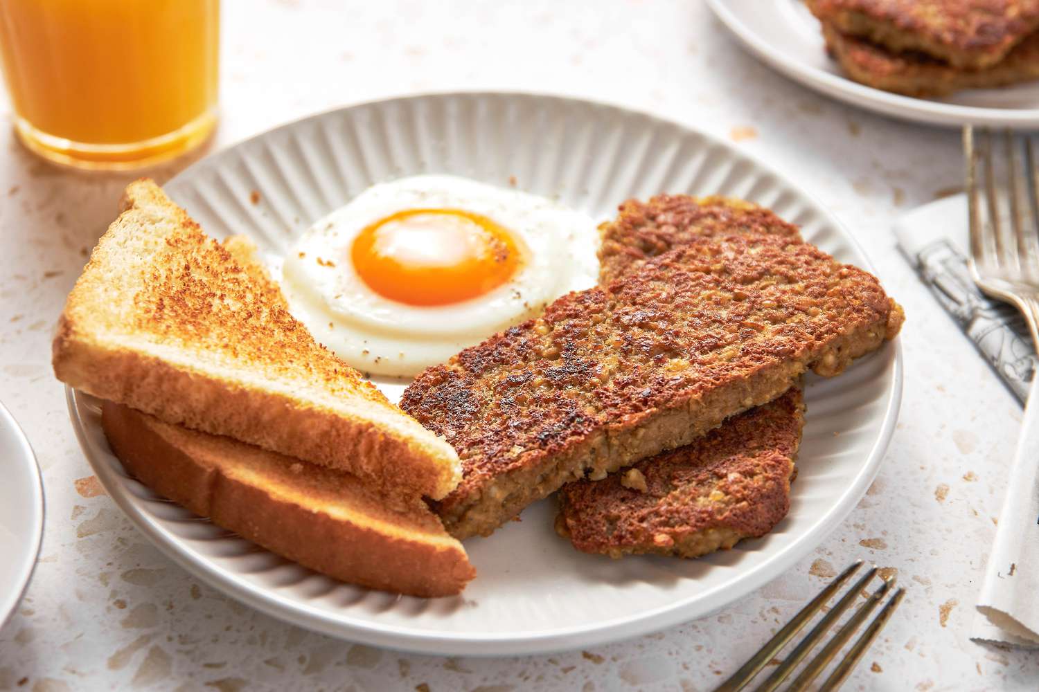 Plate With Sliced Goetta, a Sunny Side Egg, and Toast, and in the Surroundings, a Glass of Orange Juice, Another Plate With a Serving of Breakfast, a Plate With Sliced Goetta, a Plate of Toast, and Forks on the Counter
