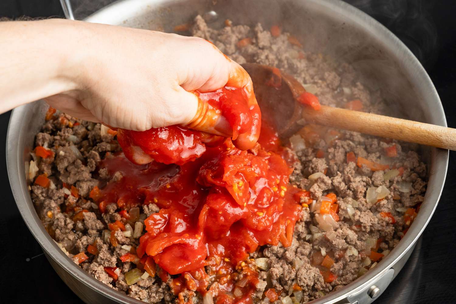 Hand crushing whole canned tomatoes into the skillet with the ground beef mixture for Johnny Marzetti casserole recipe