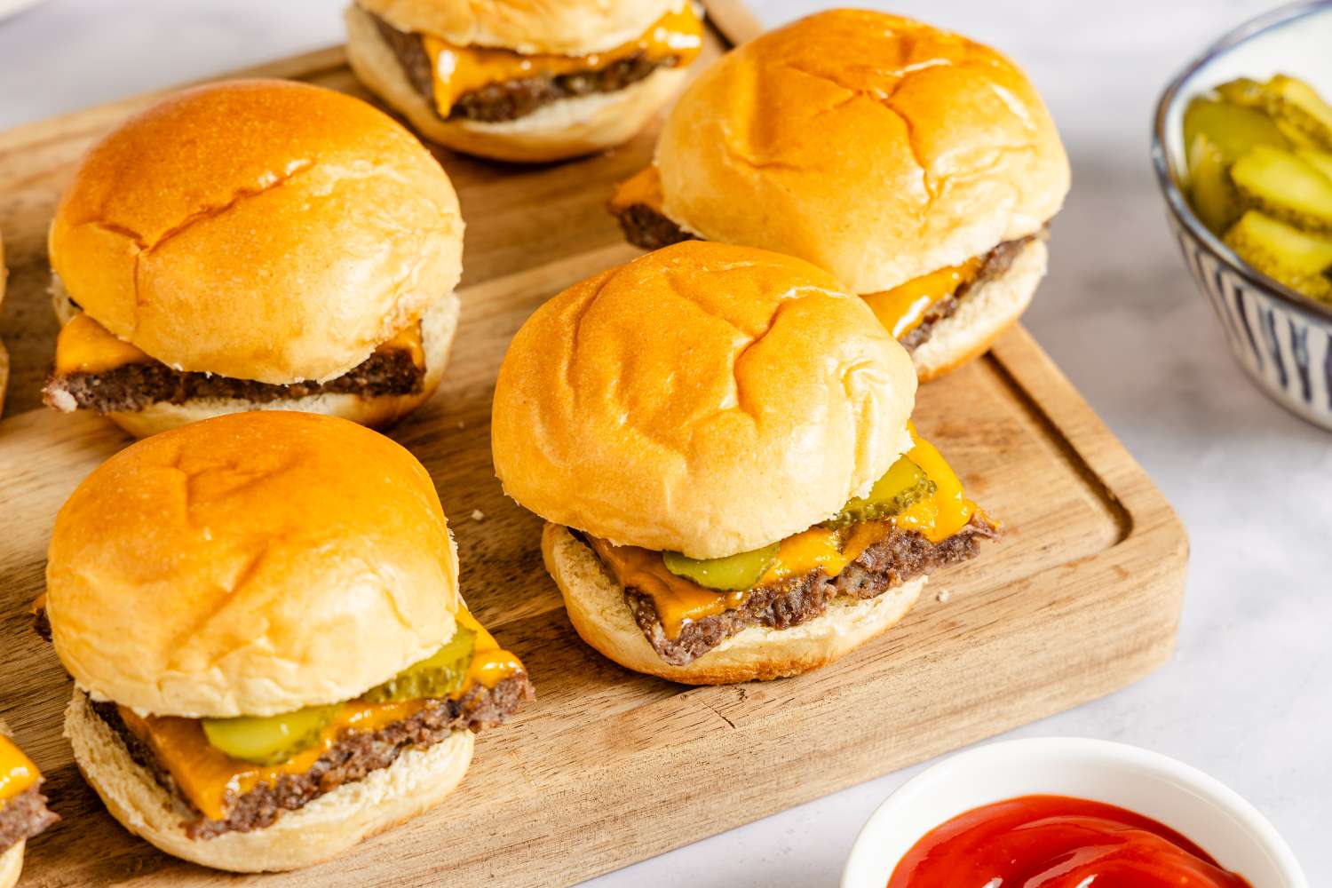 Small cheeseburger sliders on a wooden serving board with pickles and a dipping sauce bowl in the background