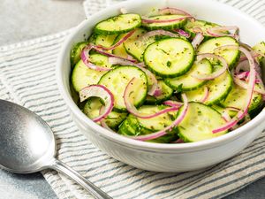 A bowl of cucumber salad with slices of cucumber and red onion on a striped cloth next to a spoon