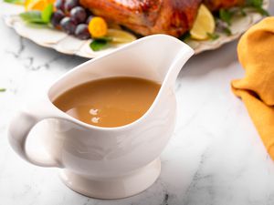 Angled view of a white gravy boat full of brown gravy next to a platter of turkey and an orange fabric napkin all on a marble counter