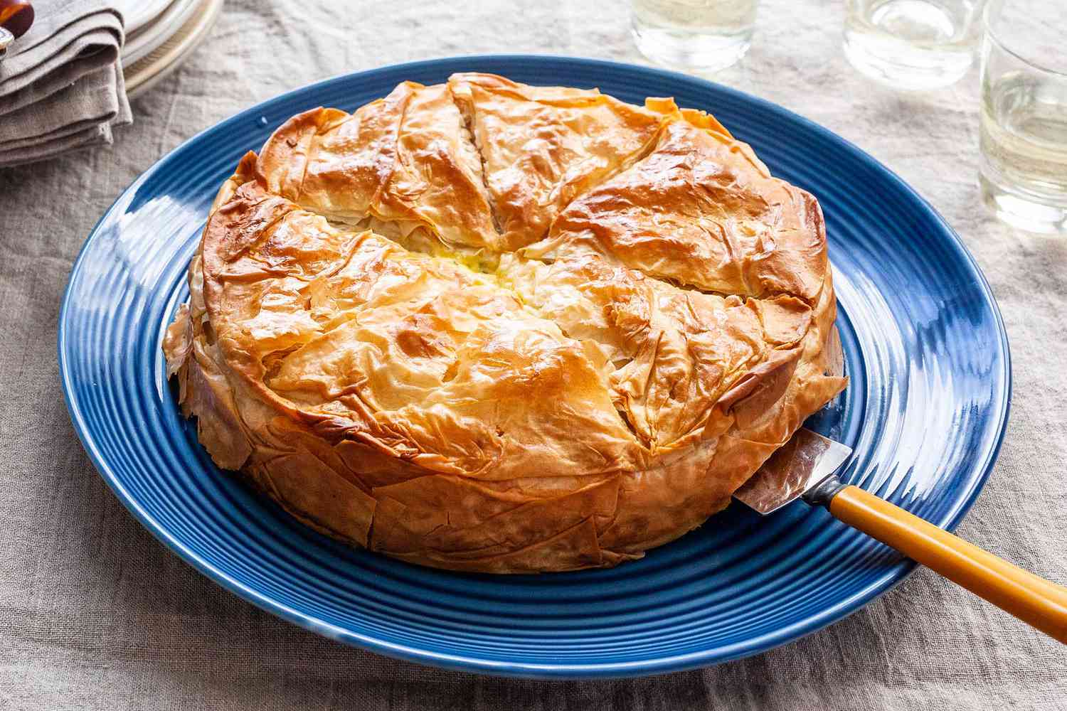 Horizontal view of a flaky cheese pie with artichokes on a blue platter. A serving utensil is under the pie. A glass is in the upper right and a stack of plates and linens is in the upper left. A grey linen covers the table.