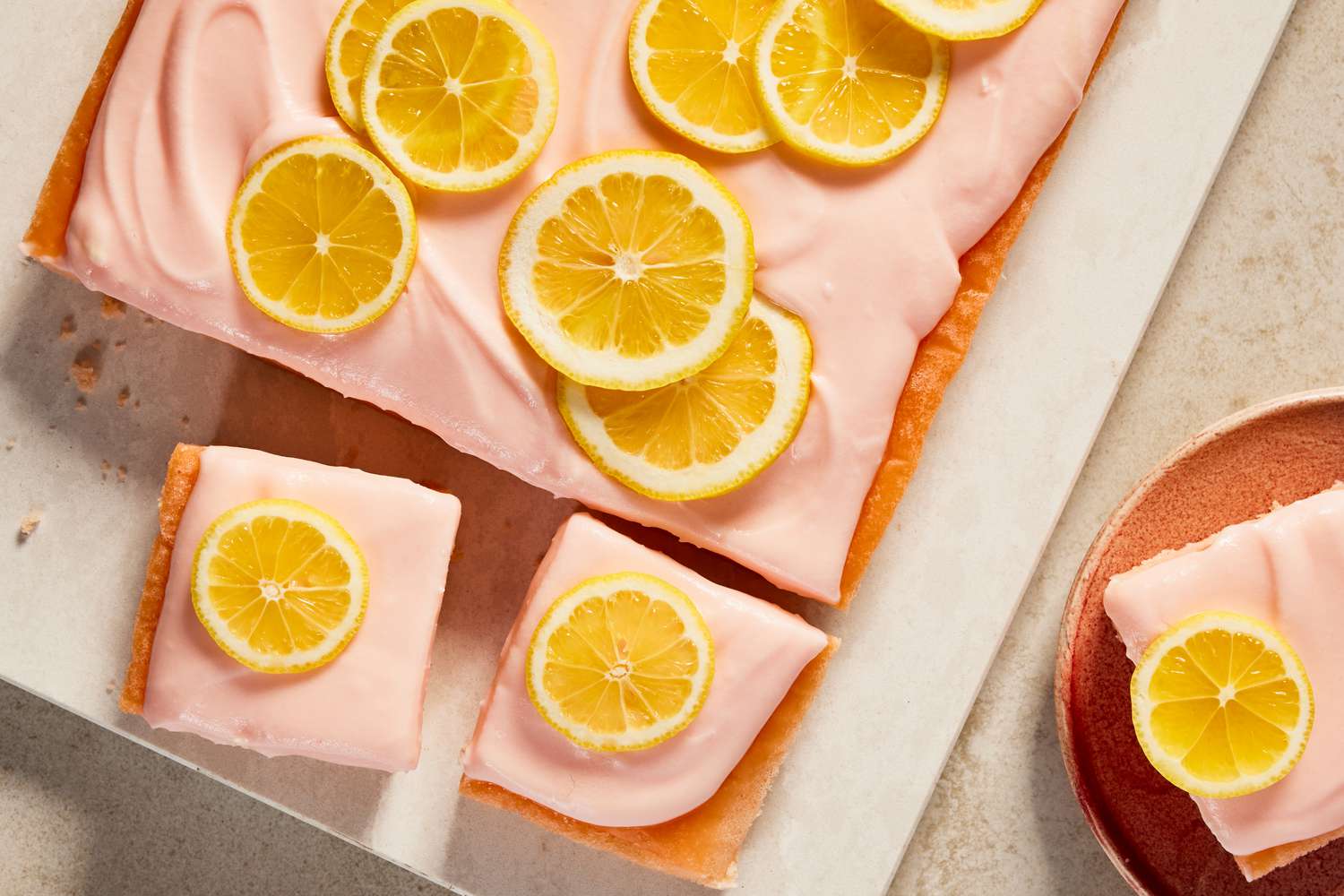 pink lemonade cake (with some slices cut out) on a board next to a plate with a slice 