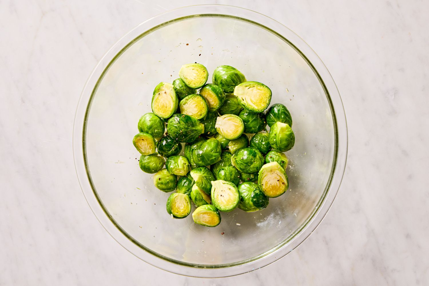 Overhead view of a clear glass bowl with sliced brussels sprouts coated with olive oil, garlic, salt, pepper, and red pepper flakes for Crispy Parmesan Brussels Sprouts Recipe 