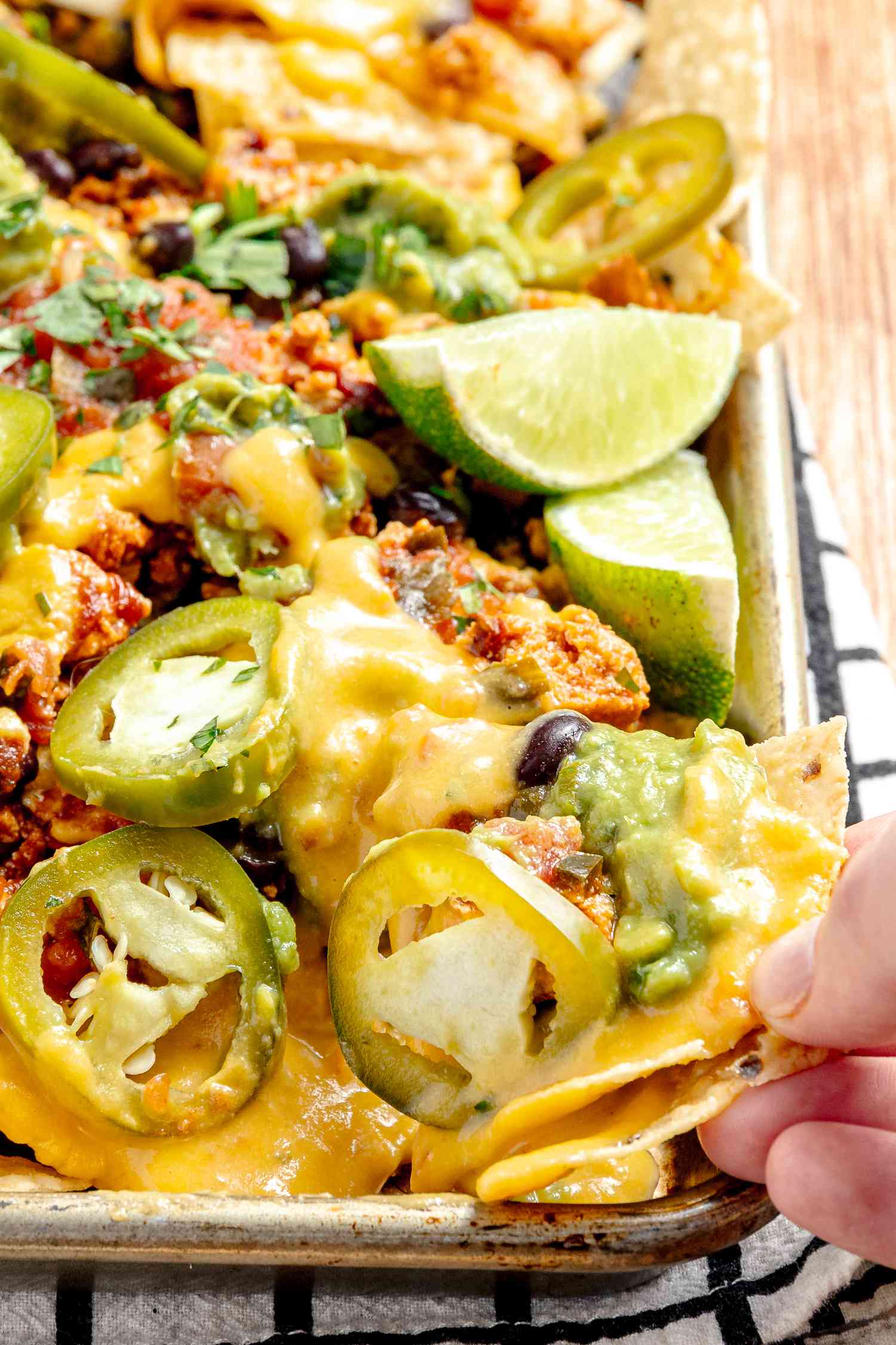 Hand Taking Some Vegan Nachos from a Baking Sheet with More Nachos (Nachos Topped with Salsa, Queso, Pickled Jalpenos, and Two Lime Wedges) 