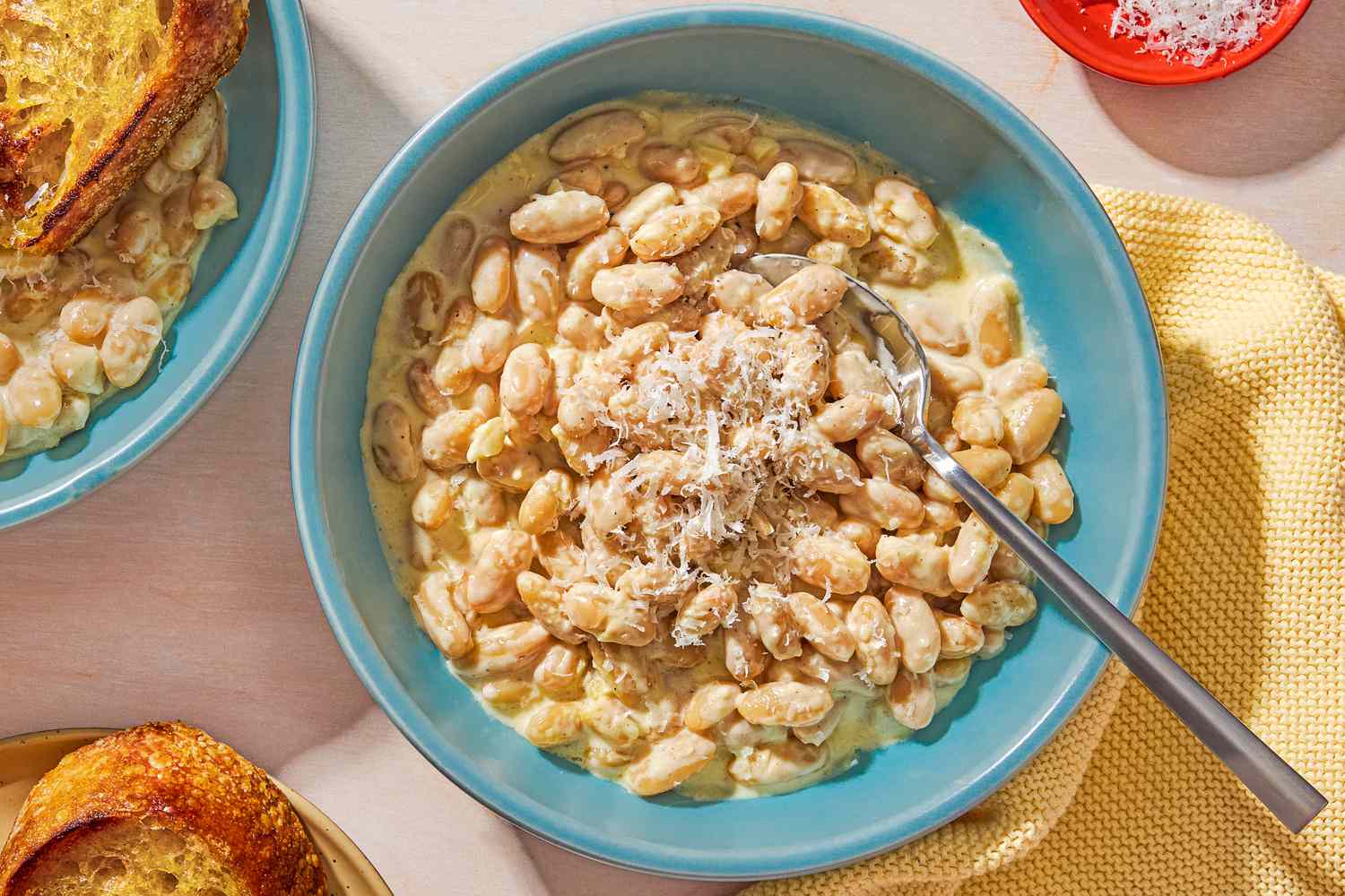 Overhead view of two teal colored bowls of 10-minute Alfredo Beans, one with a spoon and one with a piece of bread, next to a plate of bread and small red bowl of shredded Parmesan