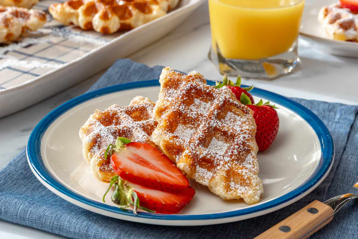 Plate with croffles strawberries powdered sugar and a glass of orange juice in the background