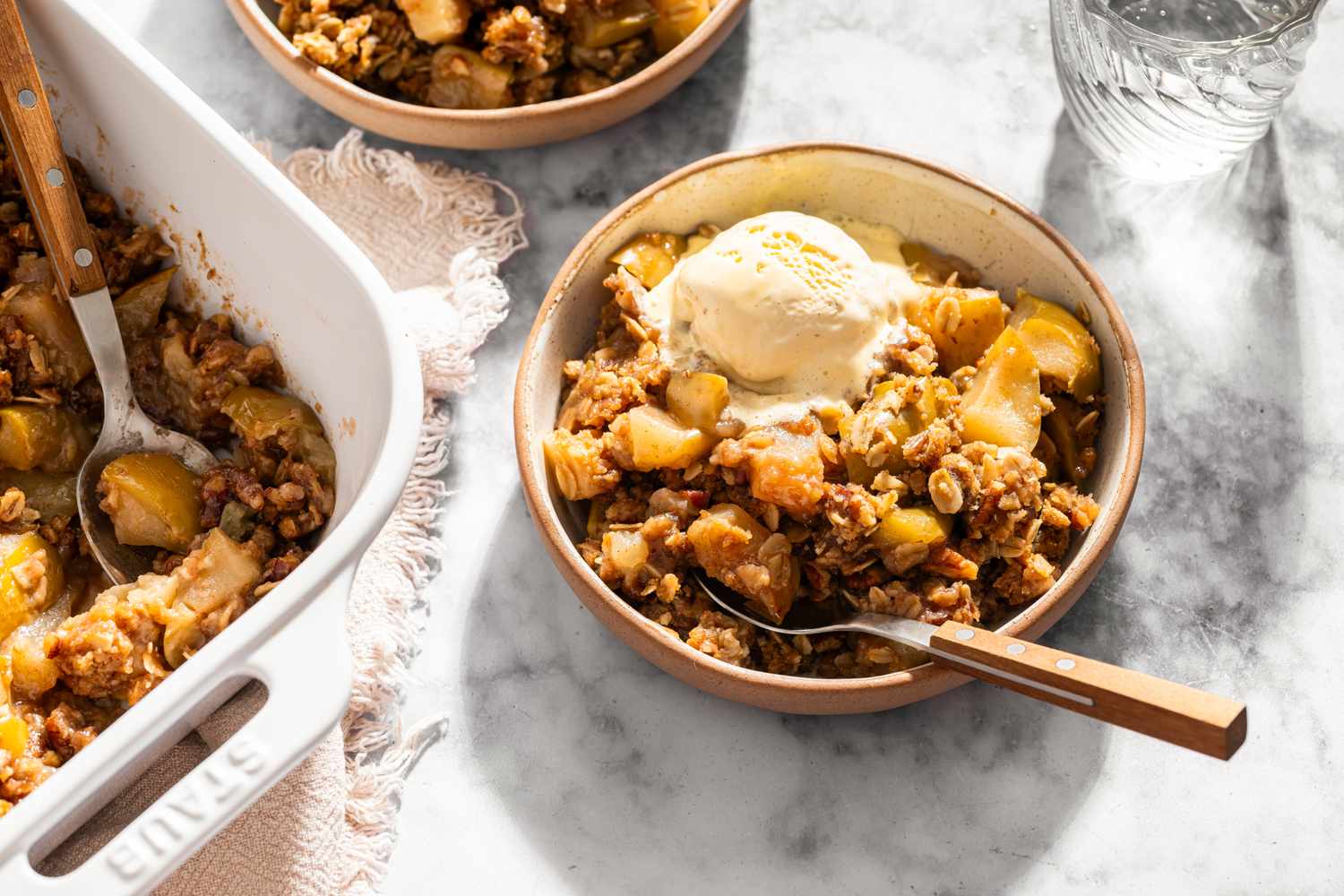 A bowl of apple crisp topped with ice cream next to a baking dish of more apple crisp
