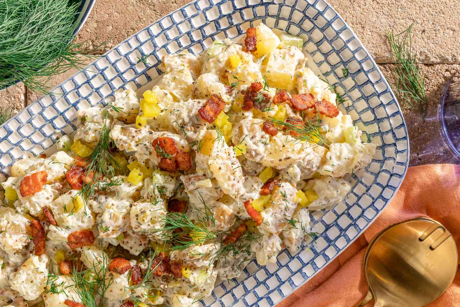 ranch potato salad on a platter next to a bowl of fresh dill and utensils on an orange kitchen towel